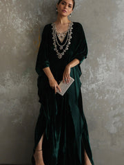 A woman models a dark emerald green velvet maxi dress with a heavily embellished silver and crystal beaded neckline, holding a silver glitter clutch against a textured gray concrete wall.