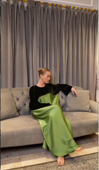 A woman sits on a gray tufted sofa in front of gray curtains, wearing a long-sleeved black and green satin dress.