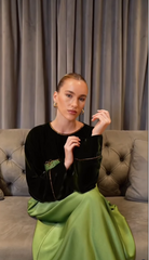 A woman seated on a tufted gray sofa wearing a black velvet long-sleeve top with gold trim and a flowing light green skirt, posed against gray curtains.