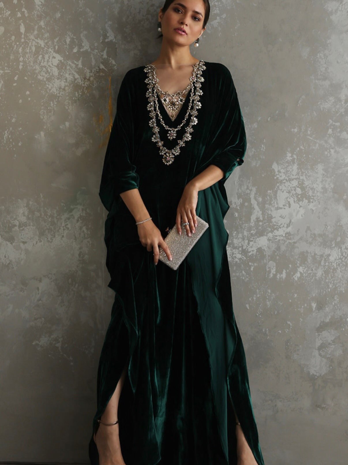 A woman models a dark emerald green velvet maxi dress with a heavily embellished silver and crystal beaded neckline, holding a silver glitter clutch against a textured gray concrete wall.