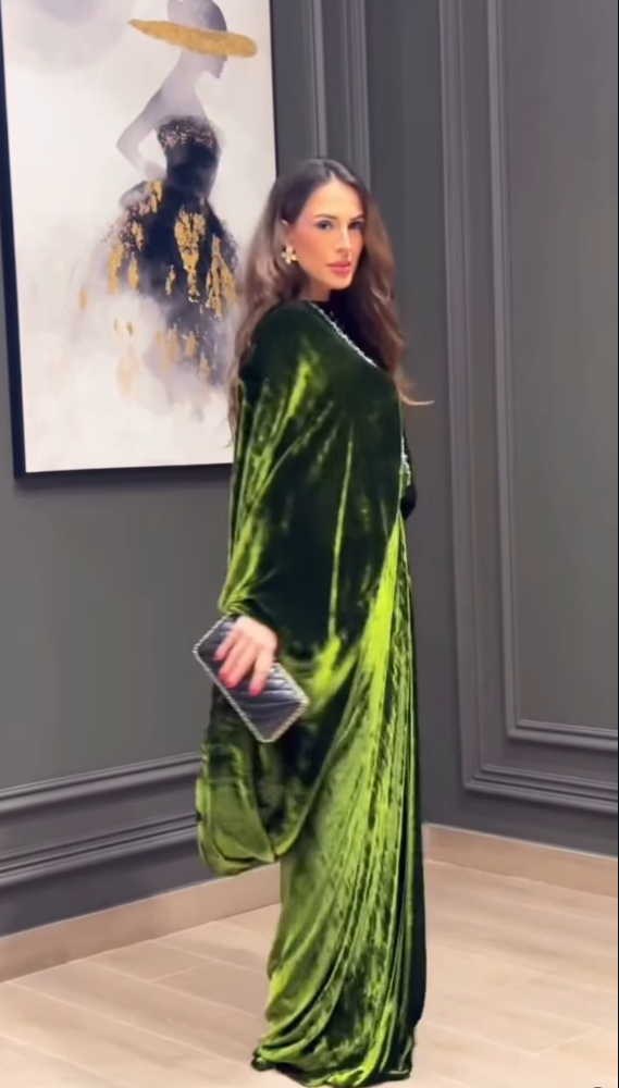 A woman wearing a floor-length emerald green velvet dress with draped sleeves and a draped hem, holding a silver clutch, standing in a room with gray paneled walls and a framed art print.