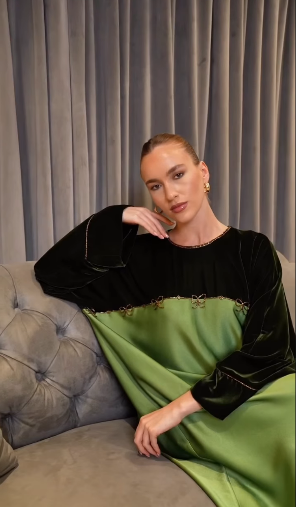 A woman wearing a long-sleeved dress with a black velvet upper section and a flowing olive green skirt, sitting on a gray tufted sofa in front of gray curtains.