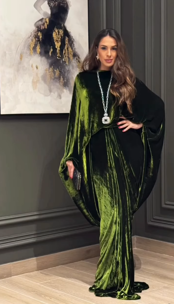 A woman models a floor-length, draped green velvet gown with wide flowing sleeves in an indoor setting with dark gray paneled walls and a framed artwork.
