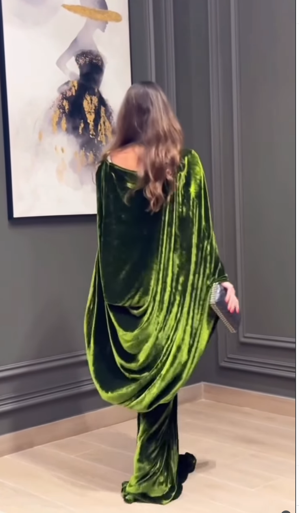 A woman stands with her back to the camera wearing a flowing olive green velvet draped dress in an indoor room with gray paneled walls and light wood flooring.