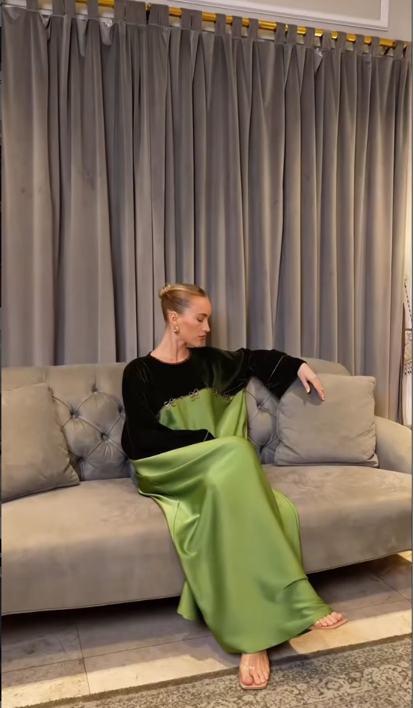 A woman sits on a gray tufted sofa in front of gray curtains, wearing a long-sleeved black and green satin dress.