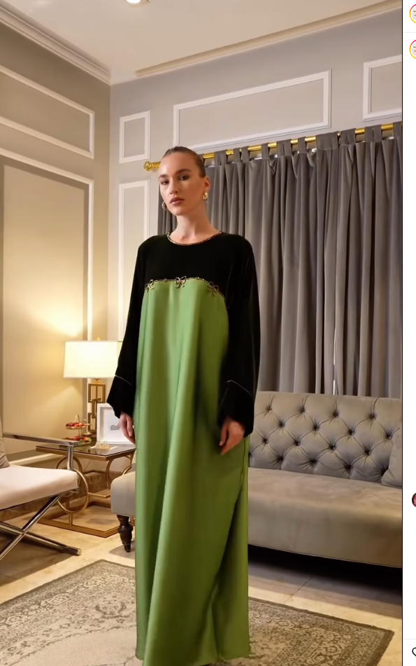 A woman modeling a long, floor-length dress in a furnished room with gray curtains, a tufted sofa, and a lamp. The dress has a black upper bodice with gold trim and a solid olive green skirt.